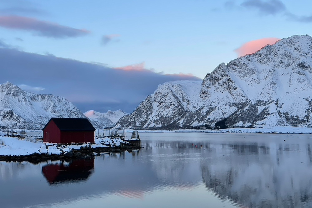 A landscape of the Lofoten Islands with snowy mountains, the Norwegian Sea and a rorbu house.