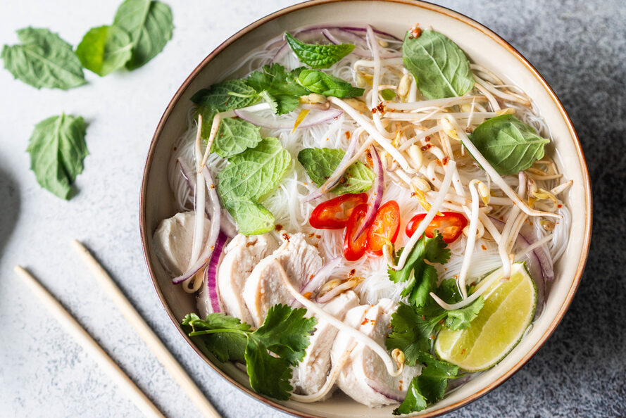 Vietnamese soup- pho ga in bowl with chicken and rice noodles, mint and cilantro, red onion, chili, bean sprouts and lime on grey background.